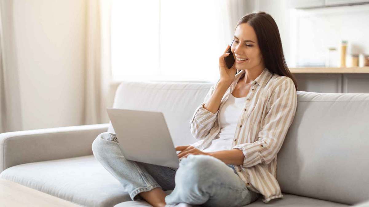 Frau sitzt zu Hause mit Laptop und Handy auf der Couch und arbeitet.