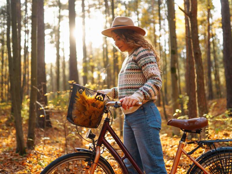 Lächelnde junge Frau in einem Hut und einem stilvollen Pullover und einem Fahrrad, genießt das Herbstwetter im Wald, unter den gelben Blättern.