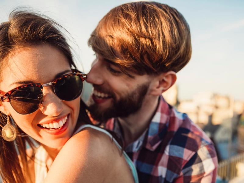 Mann umarmt Frau von hinten. Beide sitzen in der Sonne und sind glücklich.