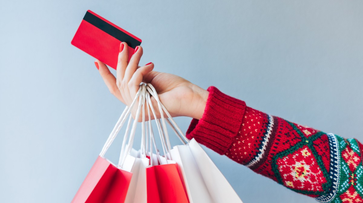 Frau mit Weihnachtspullover streckt ihren Arm raus und hält eine Kreditkarte und ein paar Tüten in der Hand.