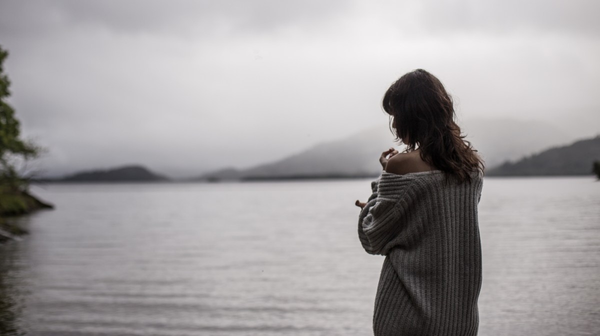 Frau mit grauem Pullover steht vor einem großen See.