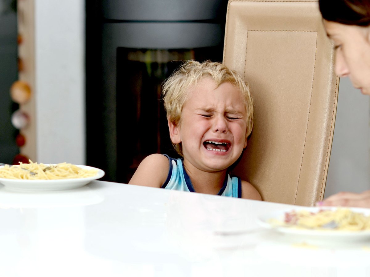 Junge sitzt mit seiner Mutter am K&uuml;chentisch und weint f&uuml;rchterlich. Die Mutter beugt sich leicht zu ihm hin&uuml;ber.