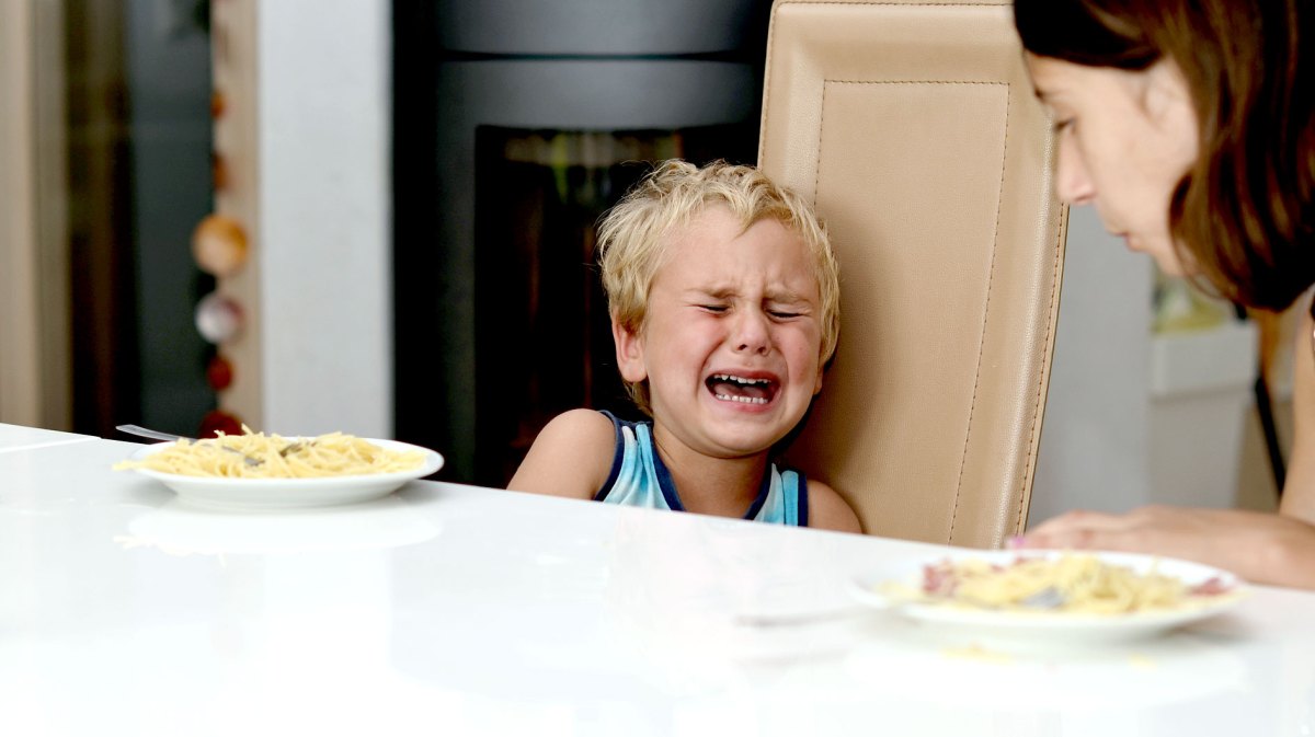 Junge sitzt mit seiner Mutter am Küchentisch und weint fürchterlich. Die Mutter beugt sich leicht zu ihm hinüber.