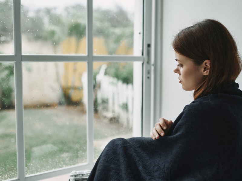 Frau, die mit einer Decke vor dem Fenster sitzt und traurig runterschaut