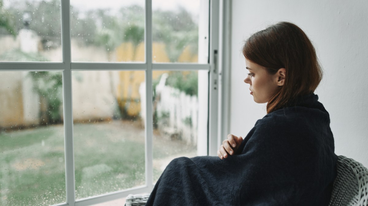 Frau, die mit einer Decke vor dem Fenster sitzt und traurig runterschaut