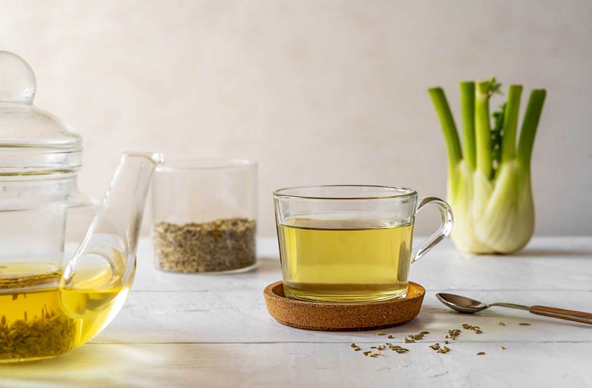 Fencheltee in einer Glastasse, frische Fenchelknolle, Samen im Glas, Teekanne auf weißem Holztisch mit hellem Hintergrund