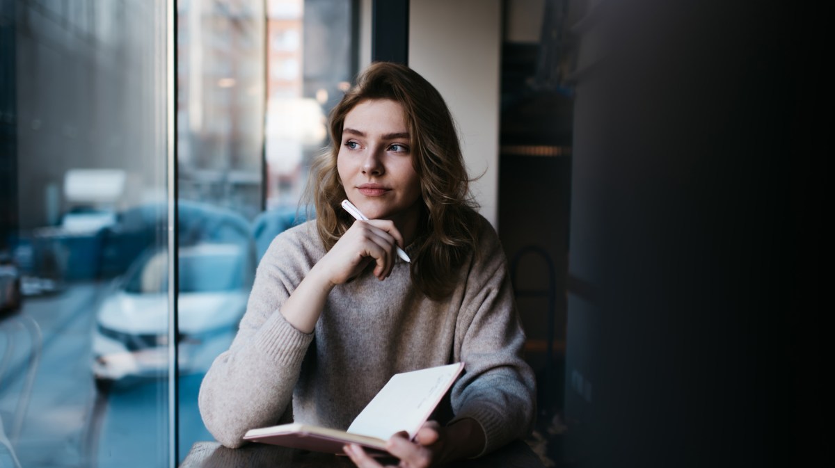 Frau vor einem Fenster, die nachdenklich hinausschaut und ein Buch mit einem Stift in der Hand hält