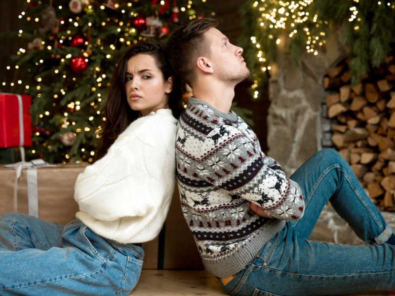 Mann und Frau sitzen Rücken an Rücken neben Weihnachtsbaum.