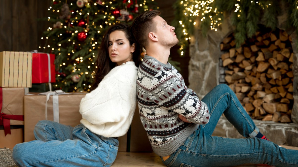 Mann und Frau sitzen Rücken an Rücken neben Weihnachtsbaum.