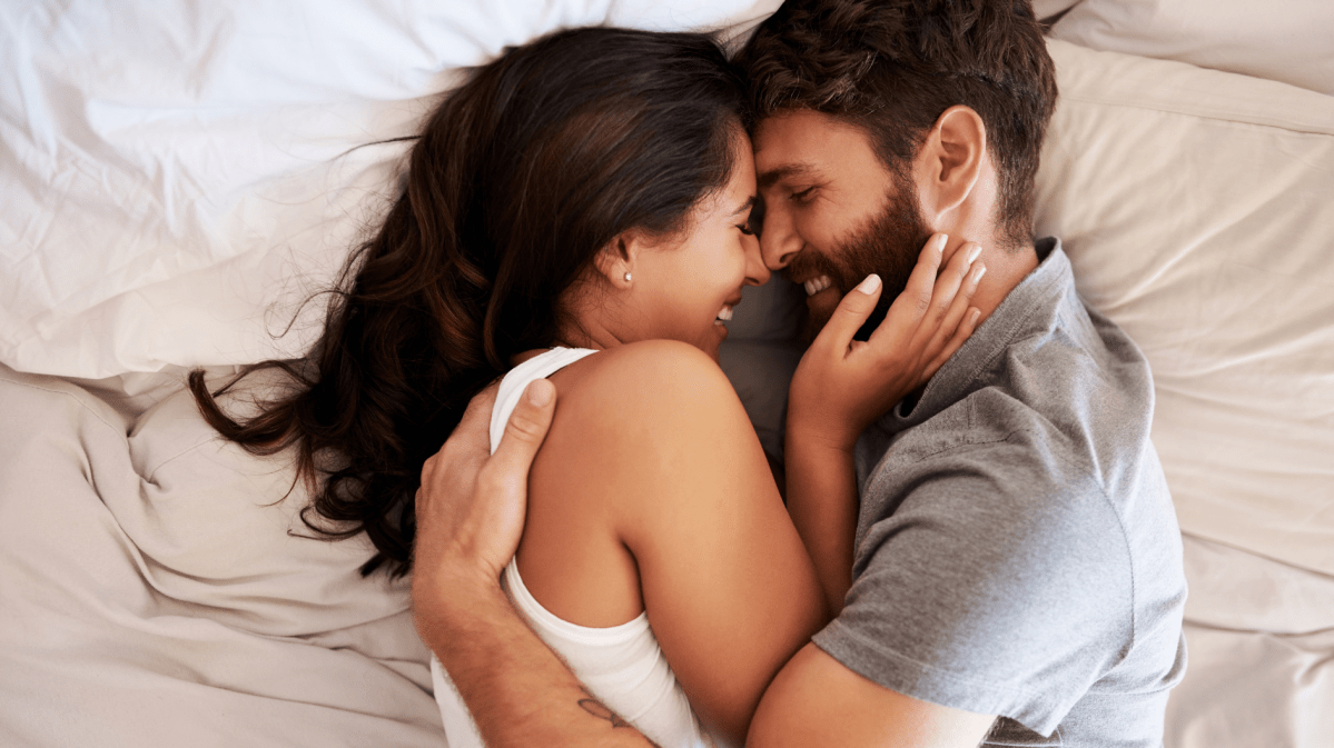 Ein Mann und eine Frau im Bett, die sich in den Armen halten, ich anlächeln und miteinander kuscheln.
