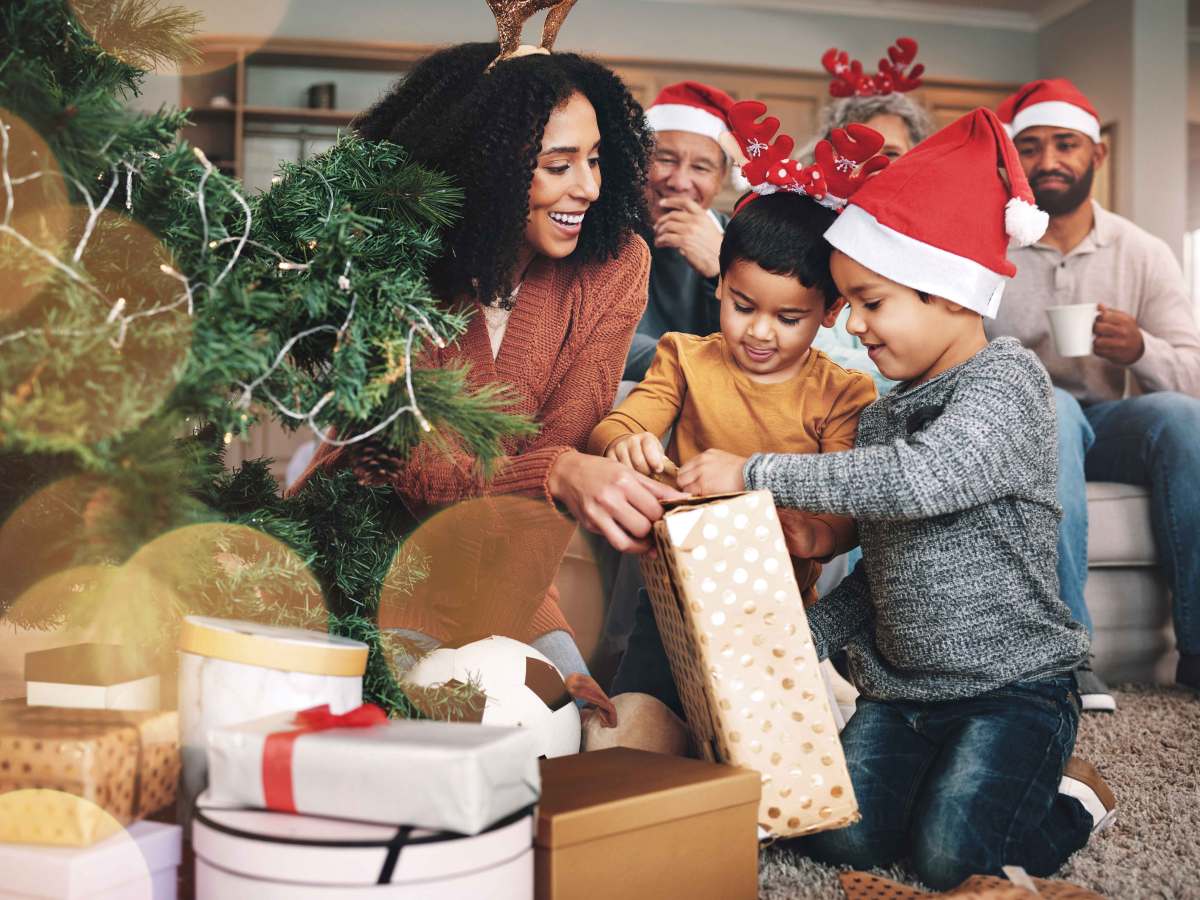 Familie sitzt am Weihnachtsbaum und die beiden Kinder reißen ein Geschenk auf.