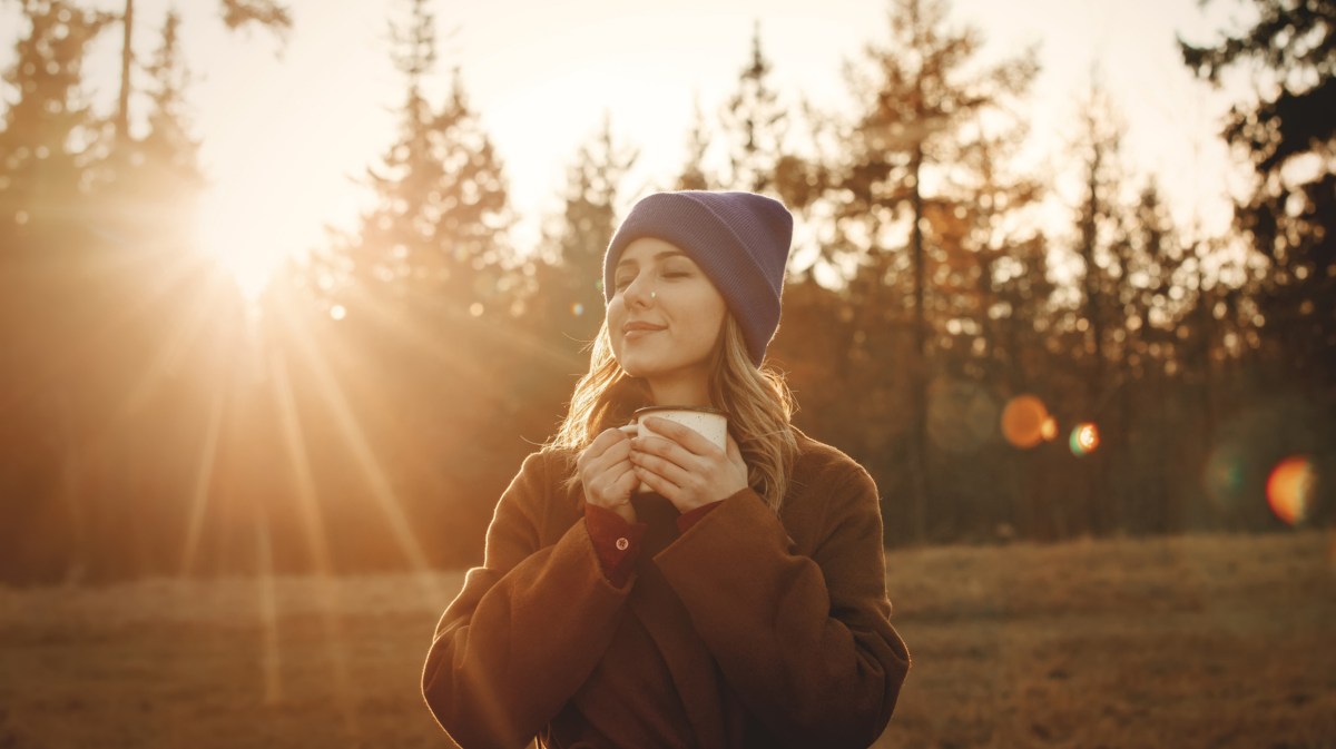 Frau im Wald mit einer Tasse in der Hand, die von Sonnenstrahlen angeleuchtet wird