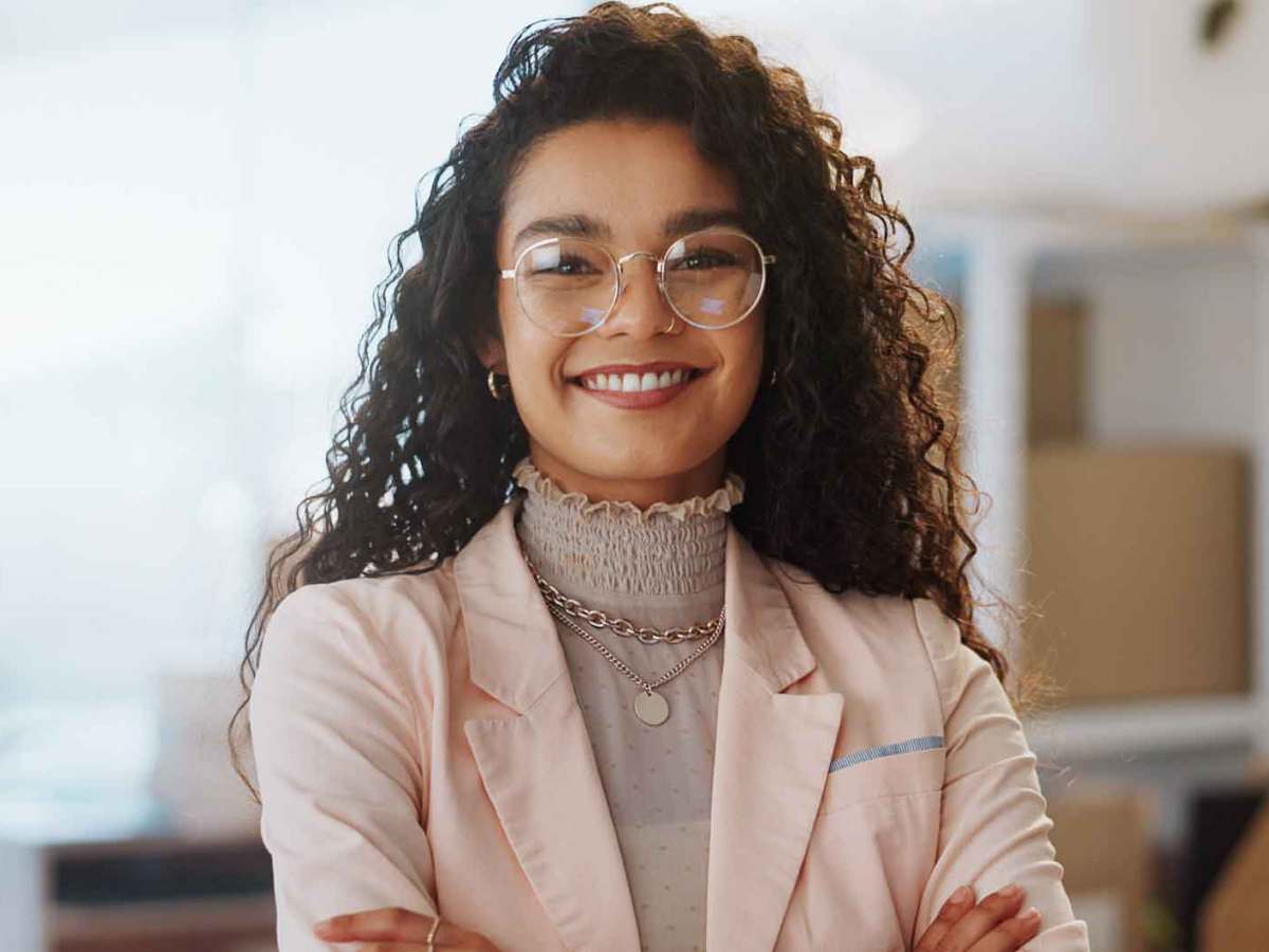 Junge Frau mit Locken und Brille lächelt selbstbewusst in die Kamera. Sie trägt ein Business-Outfit und steht auf der Arbeit.