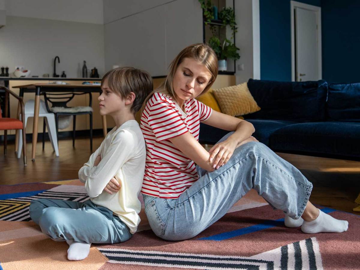 Mutter und Sohn sitzen nach einem Streit R&uuml;cken an R&uuml;cken. Der Sohn schaut starr und ver&auml;rgert in die Ferne, die Mutter schaut &uuml;ber die Schulter zu ihm.