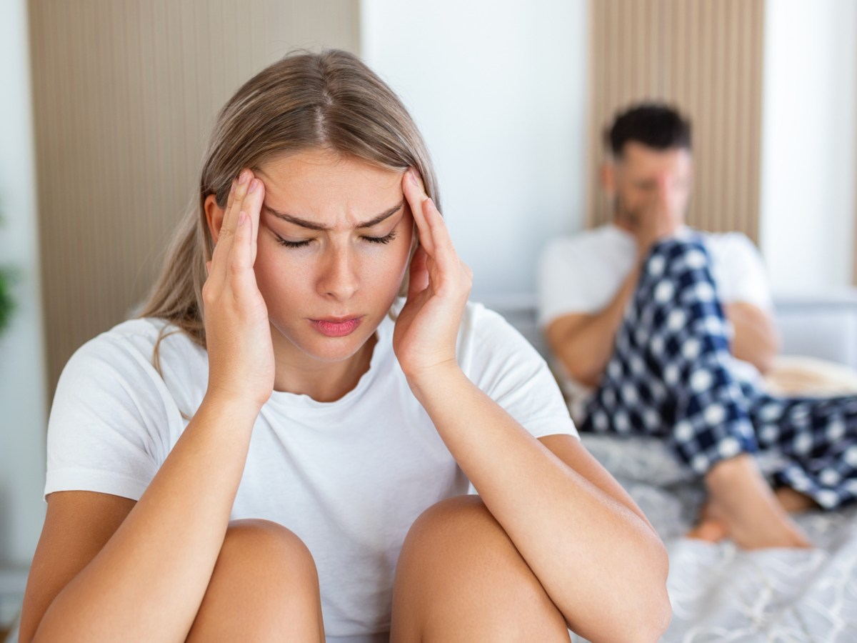 Frau auf einem Bett, die genervt ihre Hände an die Stirn packt, während im Hintergrund ein Mann auf dem Bett sitzt