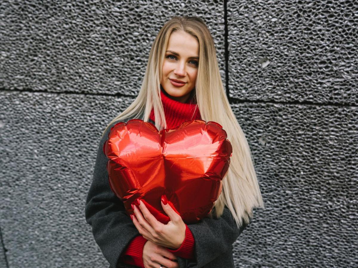 Junge Frau mit langen blonden Haaren hält einen roten Herzluftballon und lächelt sanft vor einer dunklen Steinwand.