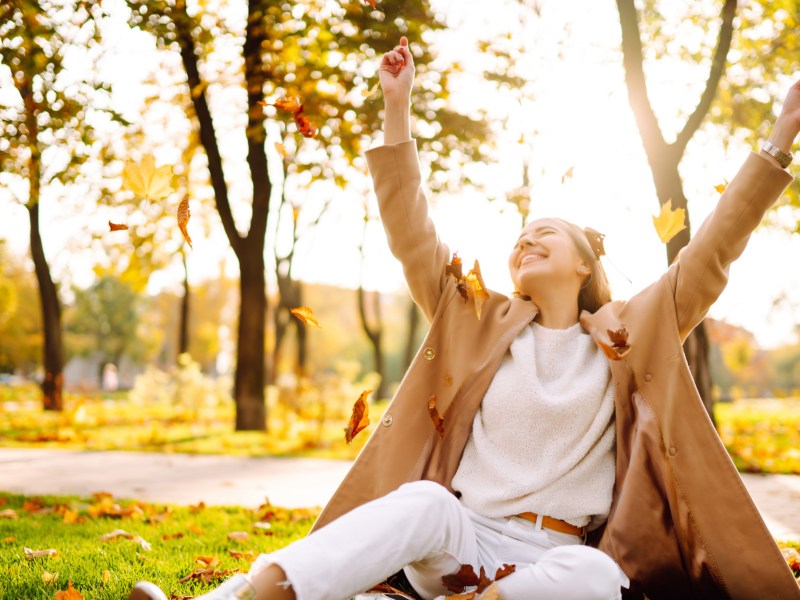 Frau im Park auf dem Rasen, die herbstliche Blätter in die Luft wirft und lacht