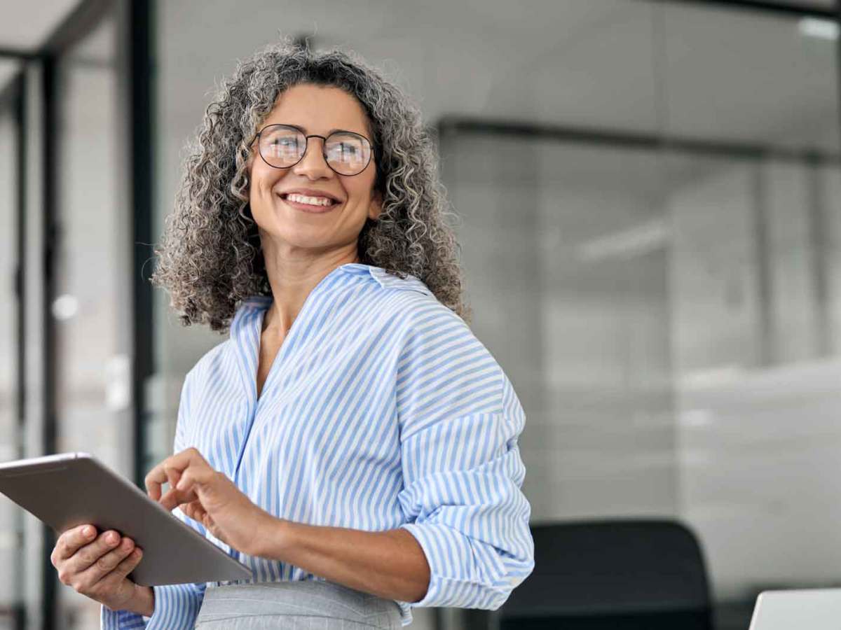 Ü50-Business Women steht auf der Arbeit mit einem Tablet und lächelt.