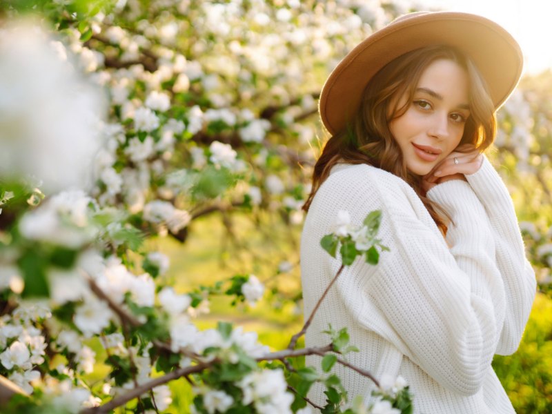 Frau mit Pullover und Hut steht vor einem blühenden Baum