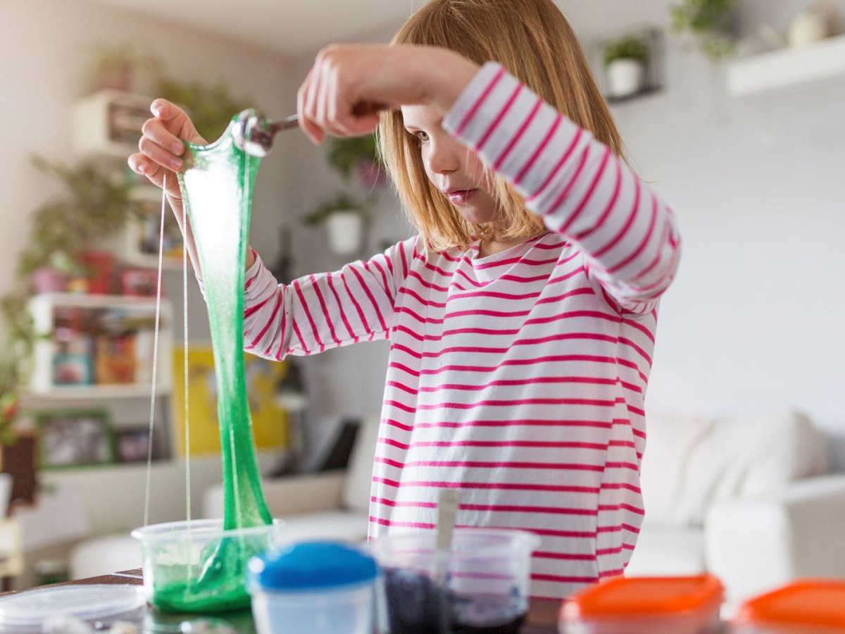 M&auml;dchen mit blonden Haaren zieht gr&uuml;nen Schleim in die H&ouml;he, umgeben von Spielzeug und Pflanzen.