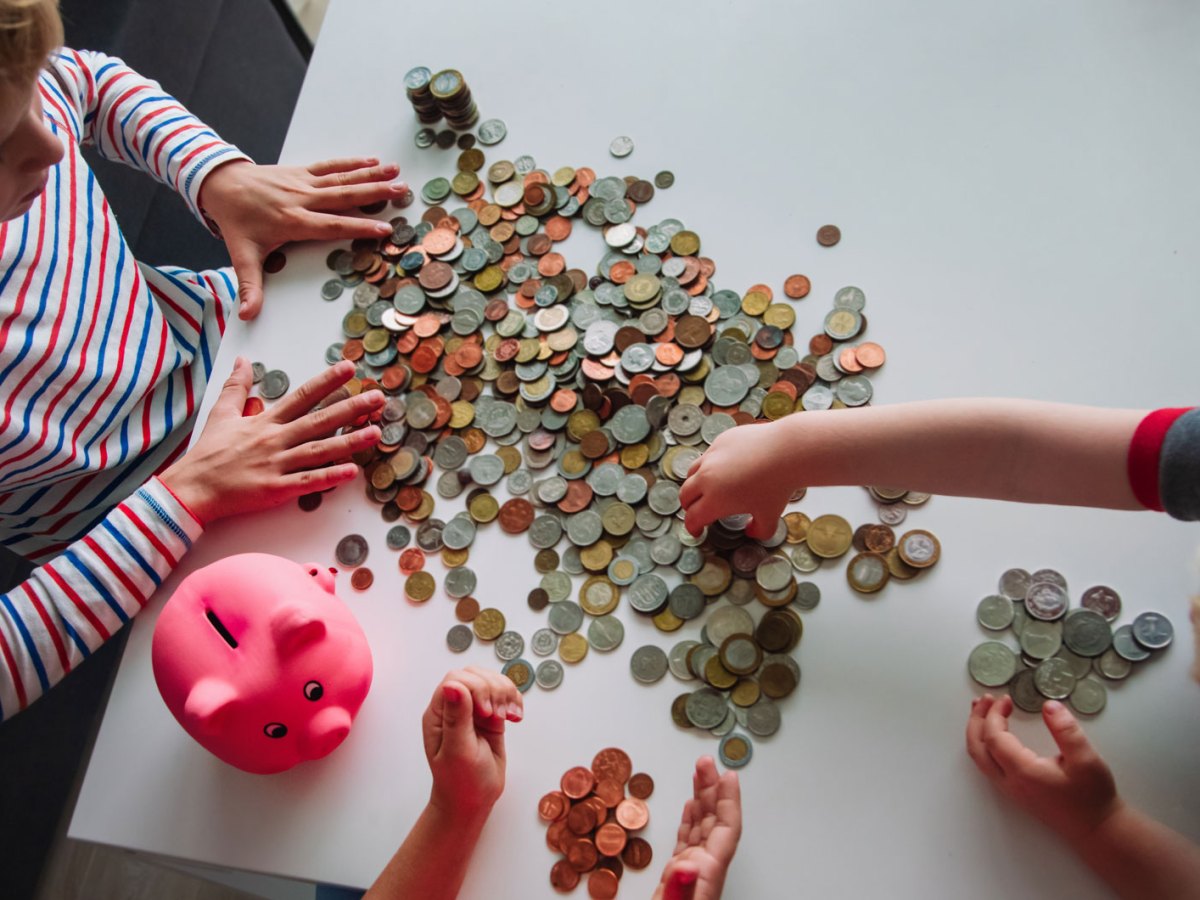 Bild von oben auf einen Tisch voller Eurom&uuml;nzen. Um den Tisch sitzen drei Kinder unterschiedlichen Alters und z&auml;hlen und sortieren das Geld.