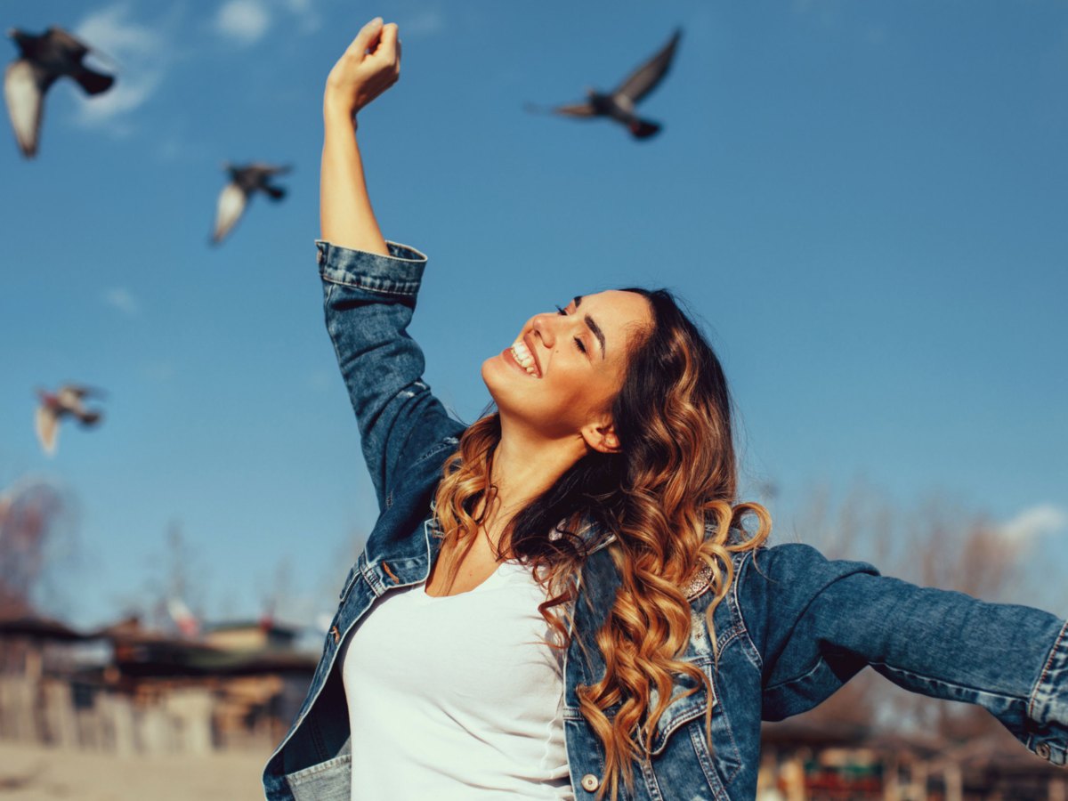 Frau mit Locken streckt die Arme in die Luft. Im Hintergrund sieht man blauen Himmel und Tauben