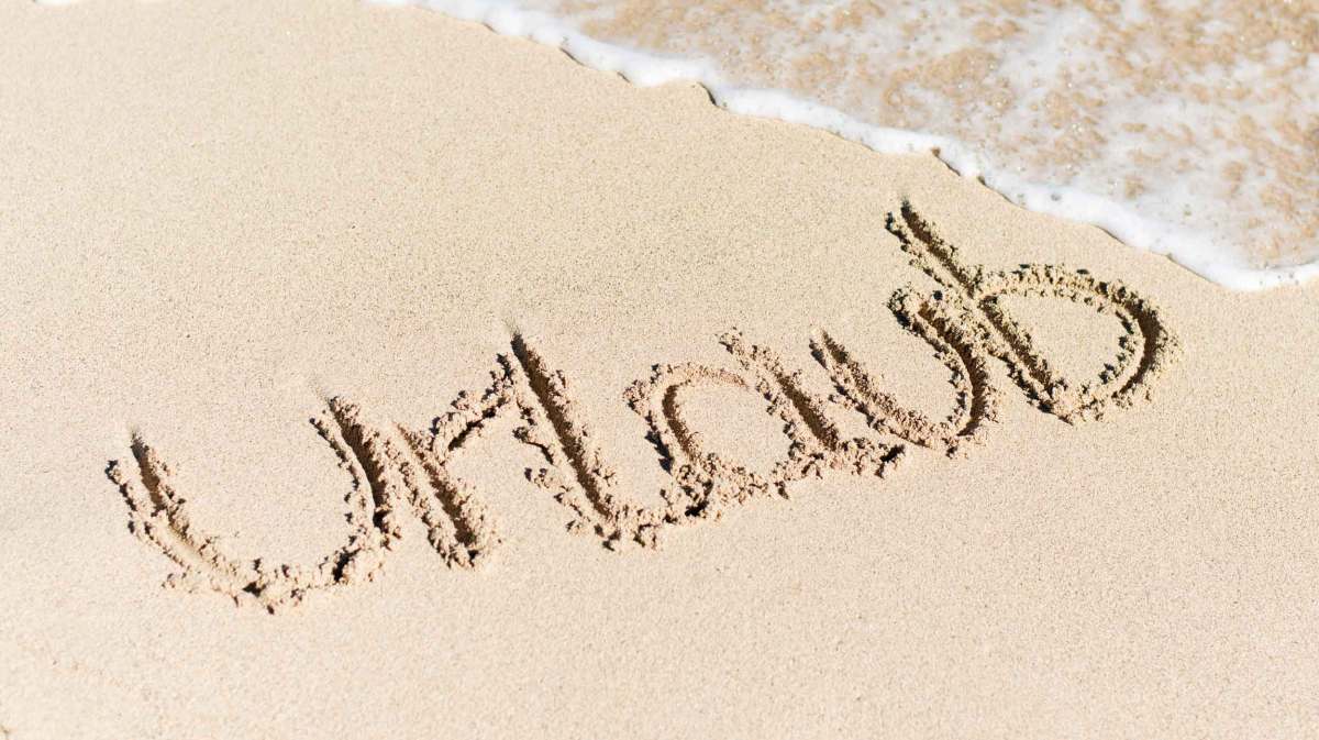 Das Wort „Urlaub“ ist in den feinen Sand eines Strandes geschrieben, während sanfte Wellen ans Ufer rollen.