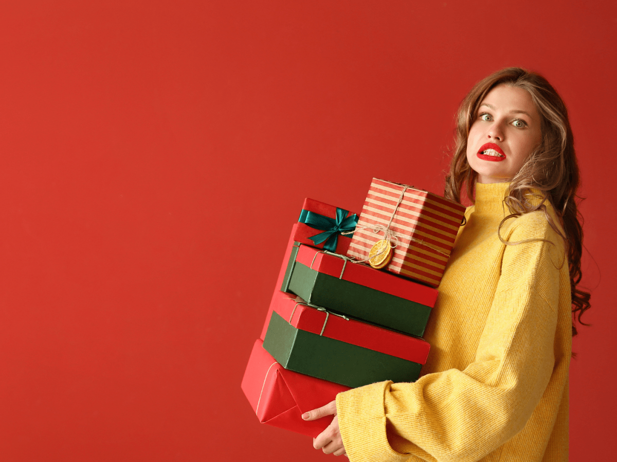 Junge Frau mit langen braunen Haaren und rotem Lippenstift tr&auml;gt stapelweise bunt verpackte Geschenke vor rotem Hintergrund.