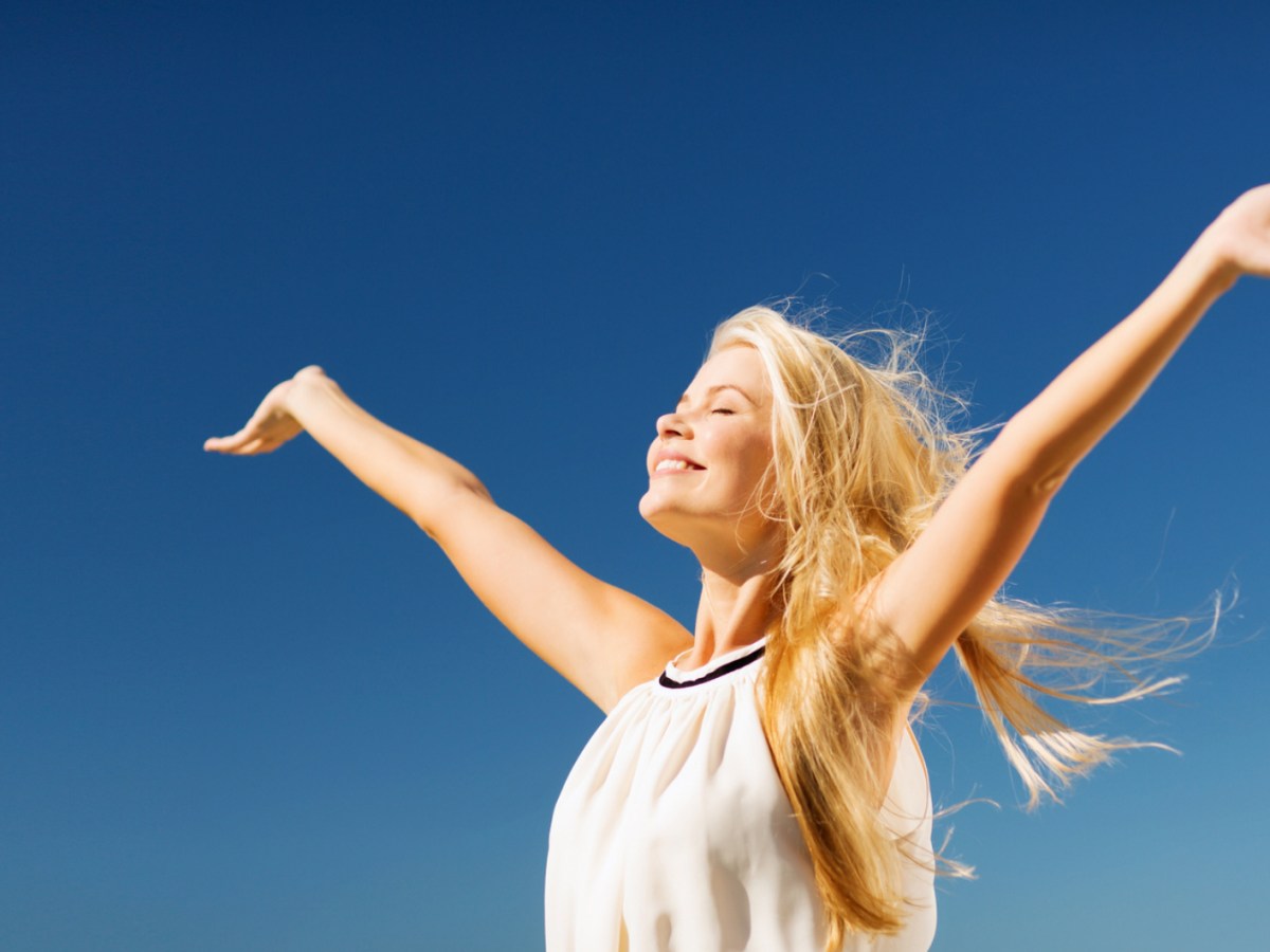 Frau im Sonnenschein vor blauem Himmel, die ihre Arme ausbreitet und in die Sonne lacht