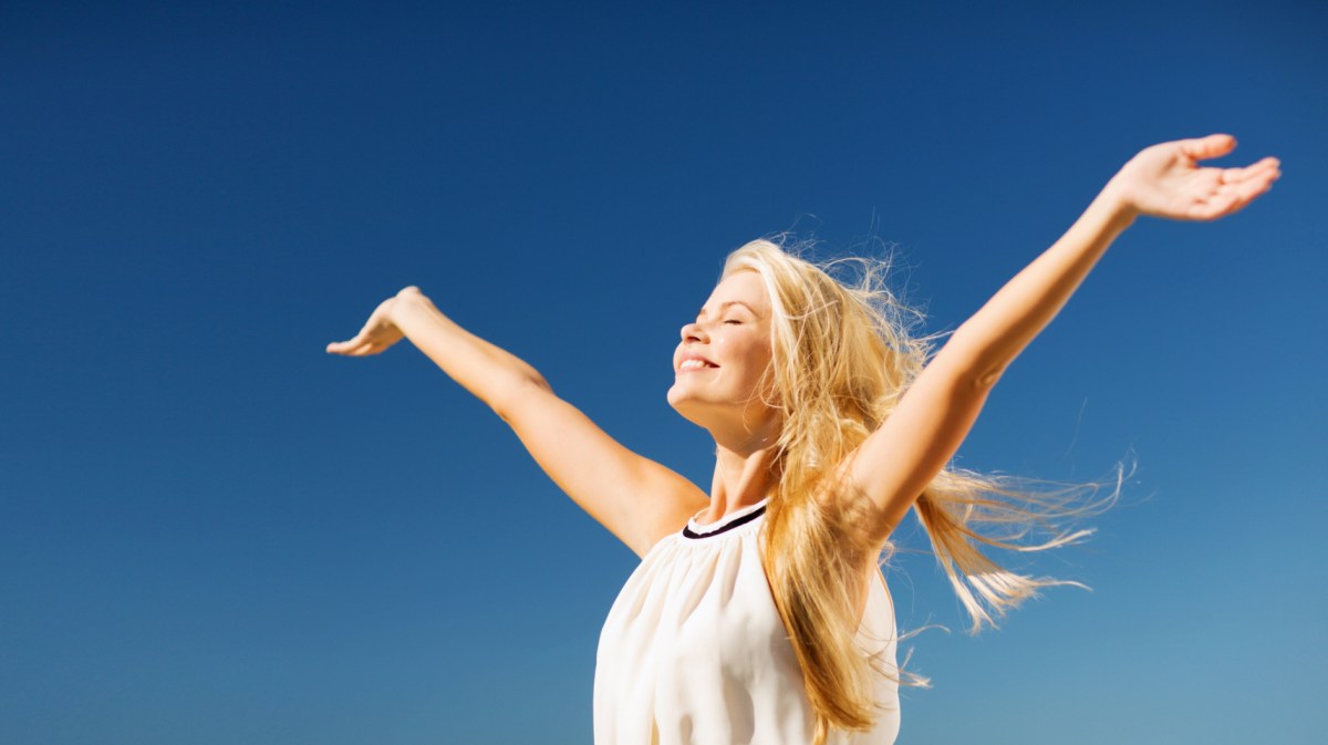 Frau im Sonnenschein vor blauem Himmel, die ihre Arme ausbreitet und in die Sonne lacht