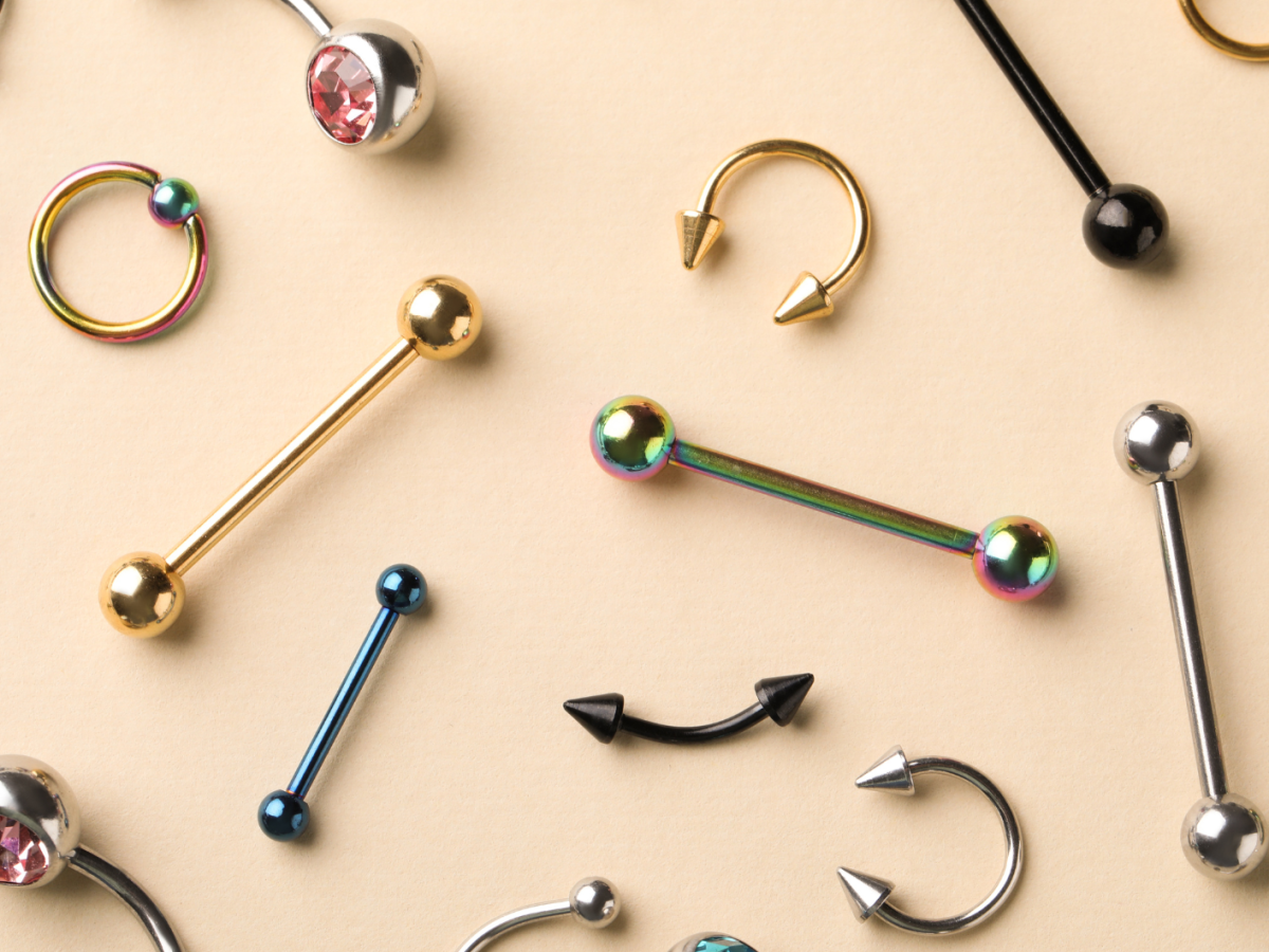 Verschiedene Piercingschmuckst&uuml;cke in Gold, Silber, Schwarz und Regenbogenfarben liegen dekorativ auf einem beigen Hintergrund.