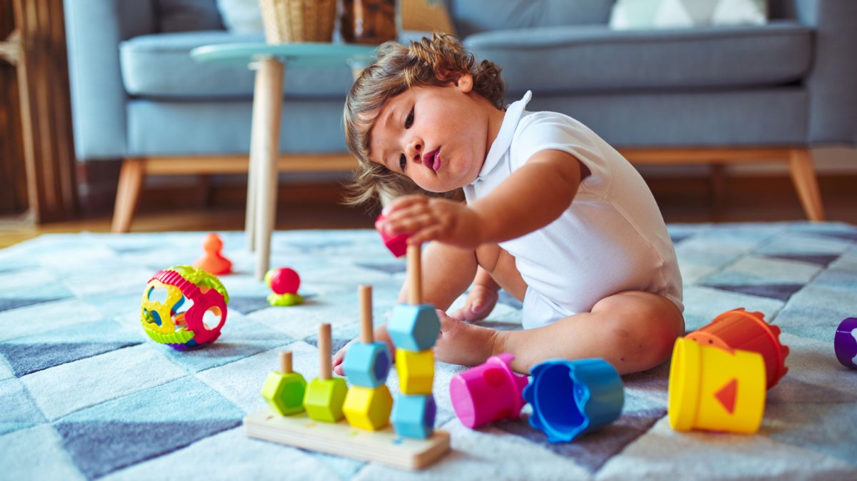 Kind, ca. zwei Jahre, sitzt auf dem Fußboden des Wohnzimmers und spielt konzentriert mit Stapelspielzeug.