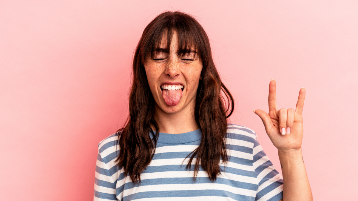 Junge Frau mit Sommersprossen und braunem Haar streckt die Zunge heraus und zeigt die Mano-Cornuta Geste vor rosa Hintergrund.