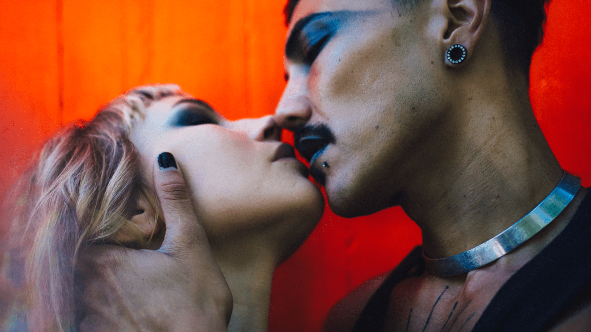 Eine blonde Frau und ein Mann mit kurzen dunklen Haaren küssen sich leidenschaftlich vor einem intensiven roten Hintergrund.