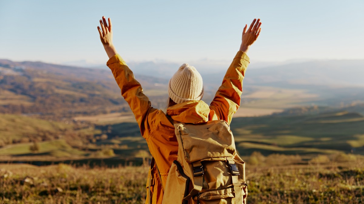 Frau vor einer Landschaft, die ihre Hände in die Luft hält und von der Sonne angeleuchtet wird