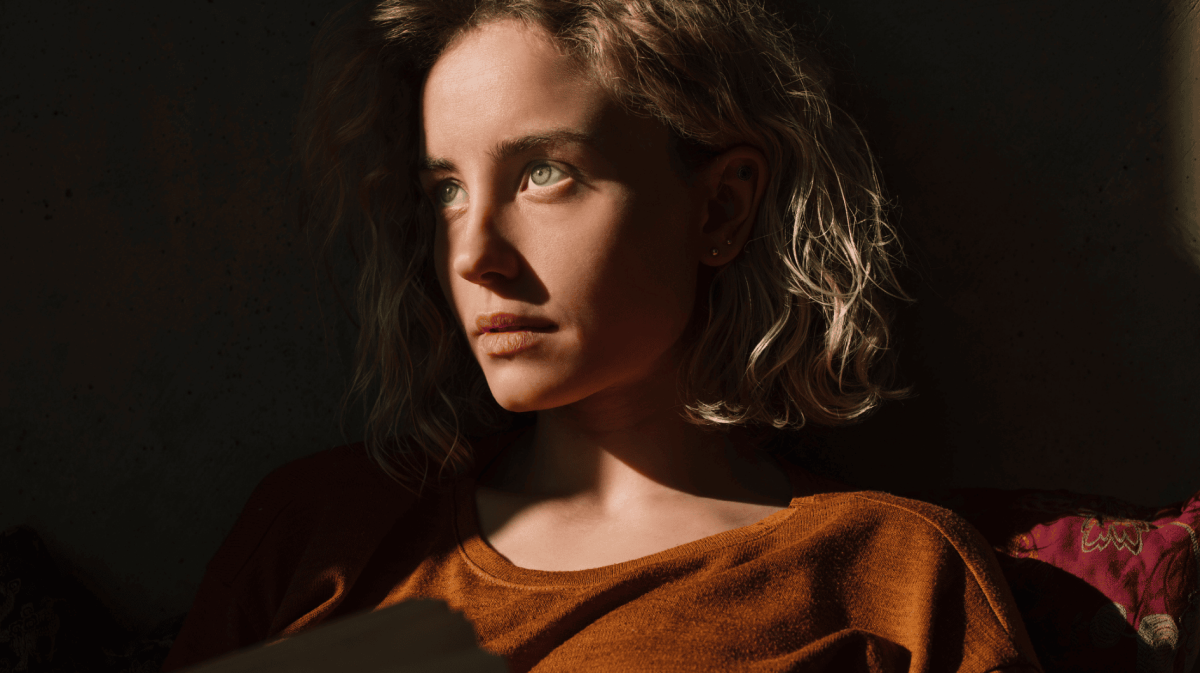 ernst blickende Frau mit orangem Pullover steht im Halbschatten, der auf ihr Gesicht fällt