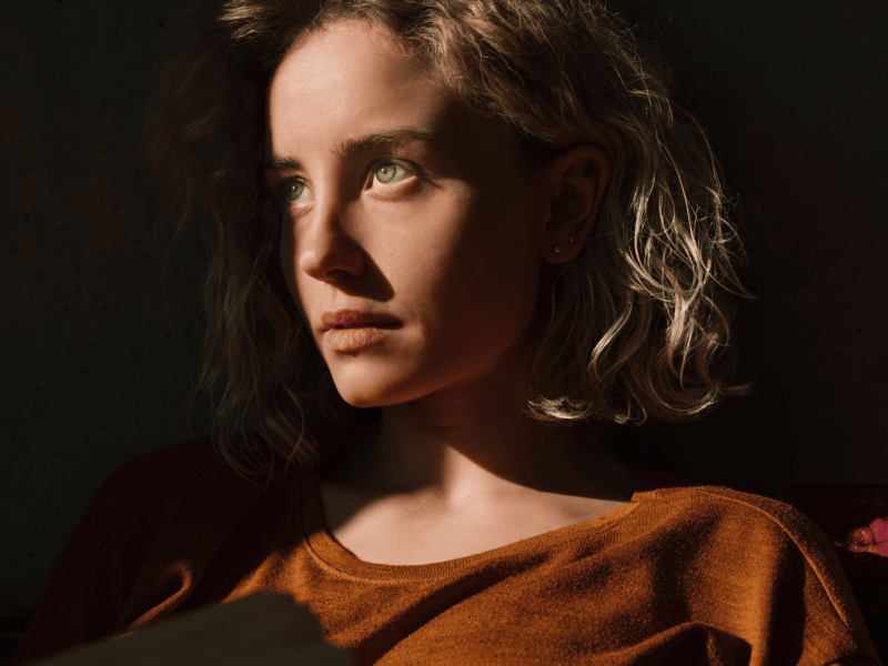 ernst blickende Frau mit orangem Pullover steht im Halbschatten, der auf ihr Gesicht fällt