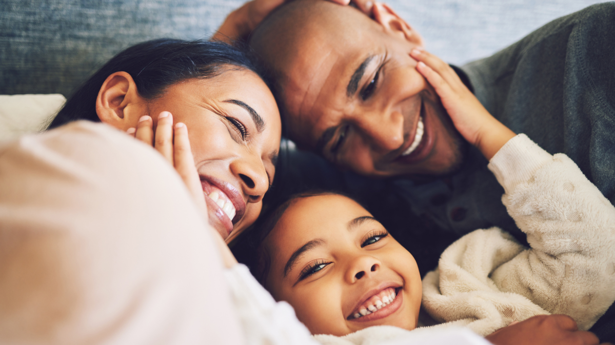 Familie, bestehend aus einer Mutter, einem Vater, einem Kind, die sich anlächelt und sich umarmt.