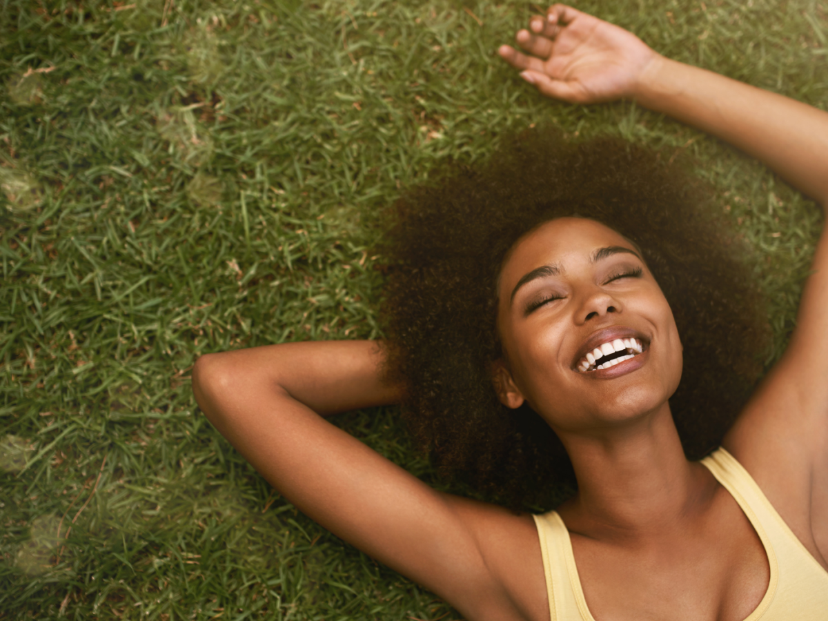 Frau auf einer gr&uuml;nen Wiese, die l&auml;chelnd in den Himmel blickt und auf ihren H&auml;nden liegt.