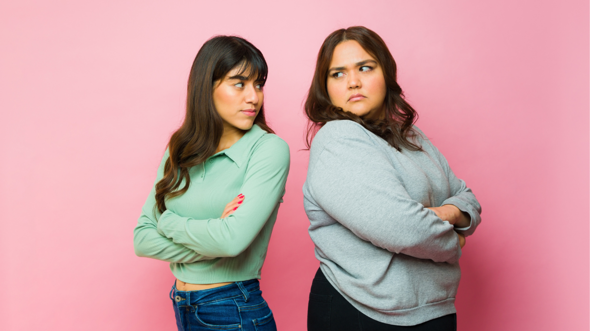 wei junge Frauen mit verschränkten Armen stehen Rücken an Rücken und schauen sich missmutig an, vor einem rosa Hintergrund.