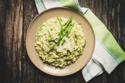 Cremiges Spargelrisotto mit geriebenem Parmesan auf einem braunen Teller, angerichtet auf einem dunklen Holztisch mit gr&uuml;n-wei&szlig;er Serviette.