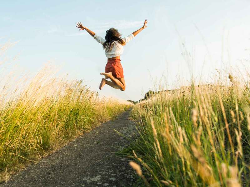 Frau springt auf einem Weg zwischen Wiesen und Feldern nach oben.