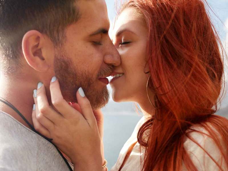 Rothaarige Frau mit langen Haaren küsst Mann mit dunklen Haaren und Bart.