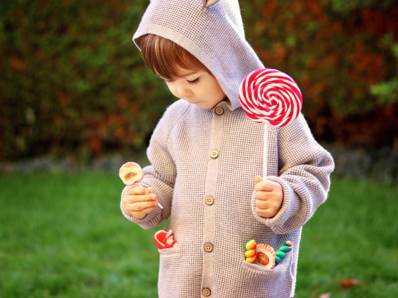 Ein Kind, ca. 4 oder 5 Jahre alt, steht draußen, mit einem großen Lolli in der Hand und weiteren kleinen in den Taschen seiner Strickjacke.