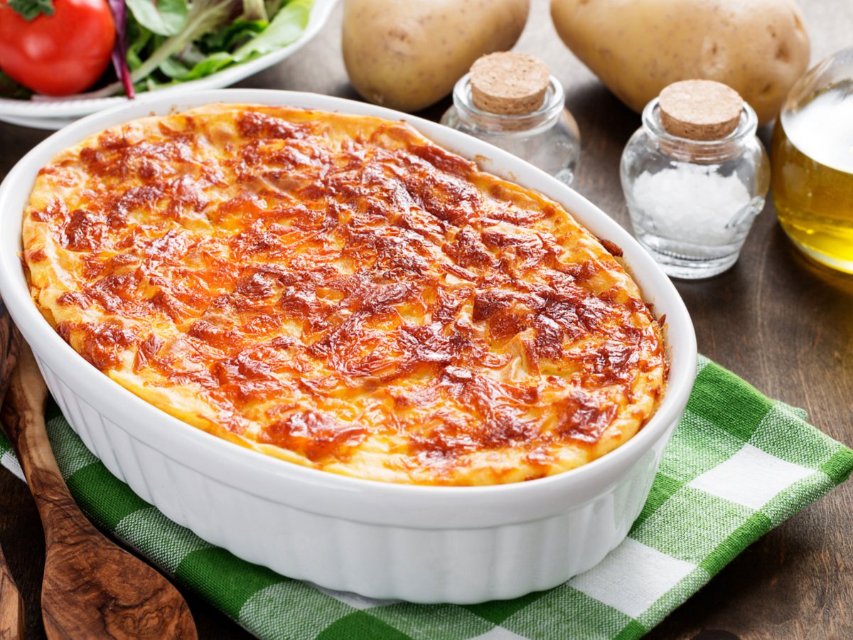 Kartoffelauflauf in wei&szlig;er Form auf Tisch, drumherum stehen verschiedene Zutaten, wie Salz, Pfeffer, Tomaten und Kartoffeln.