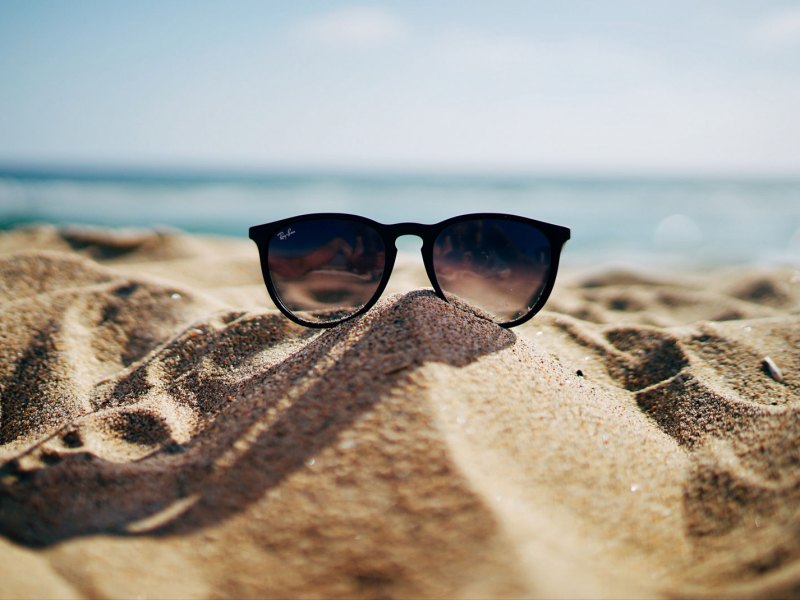 Sonnenbrille, die im Sand liegt mit Strand und Meer im Hintergrund.