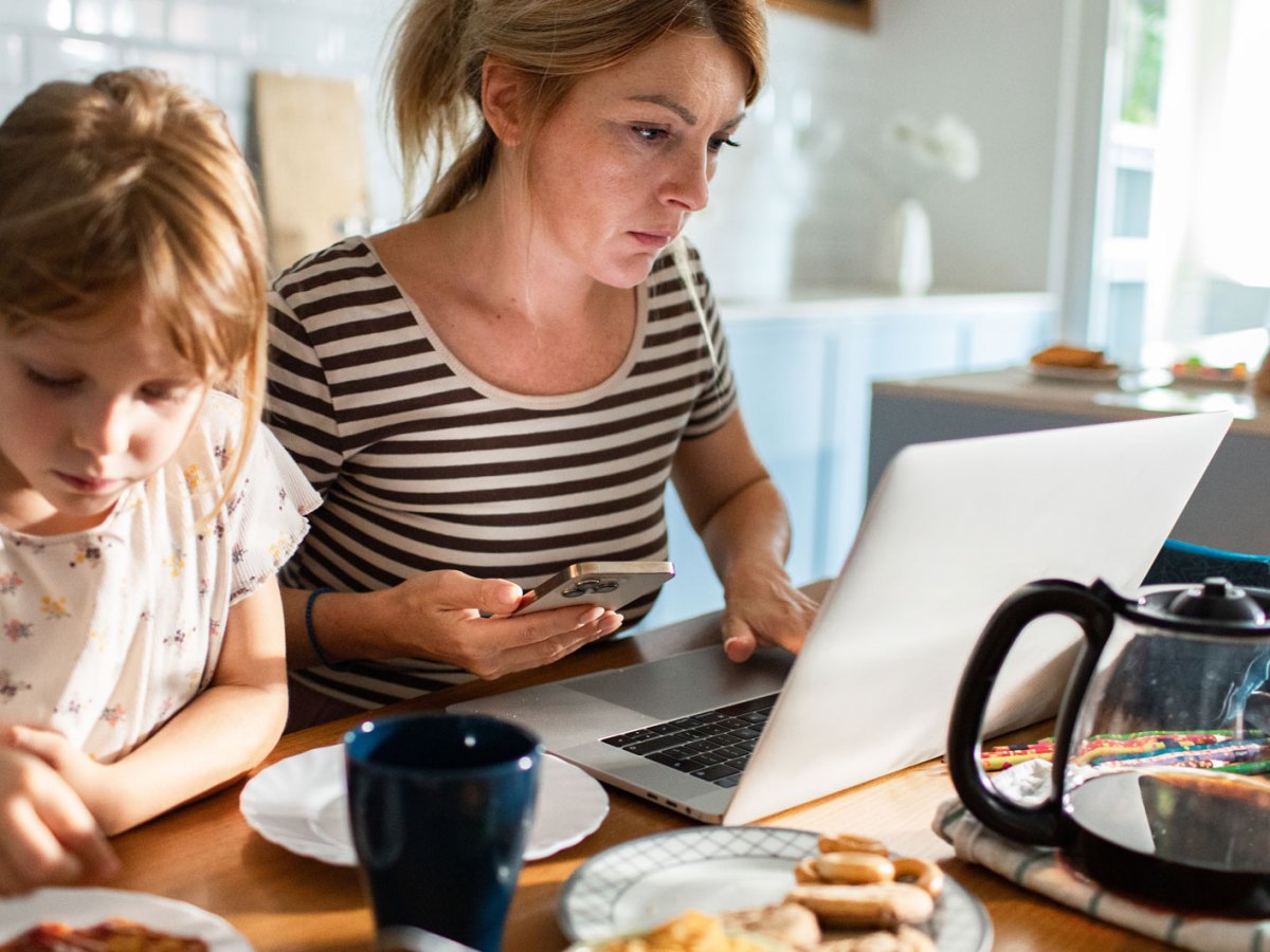 Frau sitzt w&auml;hrend des Fr&uuml;hst&uuml;cks mit der Familie am Laptop.- Vor ihr auf dem Tisch steht eine gro&szlig;e Kanne Kaffee.