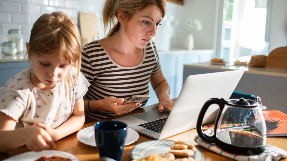Frau sitzt während des Frühstücks mit der Familie am Laptop.- Vor ihr auf dem Tisch steht eine große Kanne Kaffee.