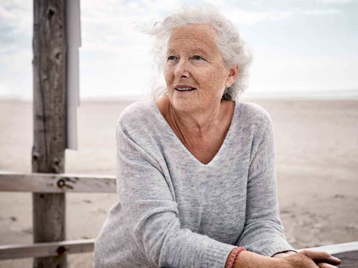 &Auml;ltere Frau steht am Strand an einen Zaun angelehnt.