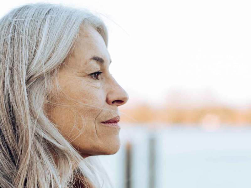 Ältere Frau mit weißen langen Haaren steht nachdenklich am Wasser.